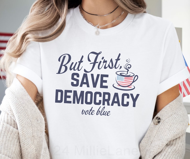 May include: White t-shirt with the text "But First, Save Democracy vote blue" in navy blue lettering. A coffee cup with an American flag design is above the text.
