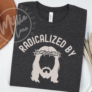 May include: A dark charcoal gray short-sleeve t-shirt featuring a light beige graphic of a figure with a beard and a crown of thorns. The text RADICALIZED BY is printed in a curved beige font above the graphic. The shirt has a crew neckline.