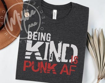 以下が含まれることがあります： 白と赤で「BEING KIND IS PUNK AF」の文字が入った、ダークグレーのクルーネックTシャツ。柔らかい素材でできており、カジュアルウェアに適しています。背景には金のネックレスと茶色の帽子があります。