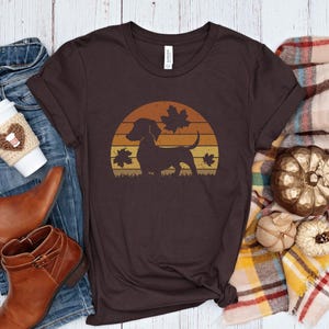 May include: A brown t-shirt with a dachshund silhouette set against a sunset design and autumn leaves. The shirt is styled with blue jeans, brown boots, and a coffee cup with a heart-shaped cosy, evoking an autumnal theme.