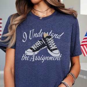 Könnte beinhalten: Ein marineblaues T-Shirt mit dem Text "I Understand the Assignment" und einer Grafik von zwei schwarzen und weißen Turnschuhen.