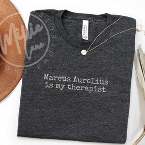 以下が含まれることがあります： 「Marcus Aurelius is my therapist」の白文字がプリントされた濃いグレーのTシャツ。丸いペンダントが付いたゴールドカラーのネックレスがシャツの上にあります。茶色の帽子が一部見えています。