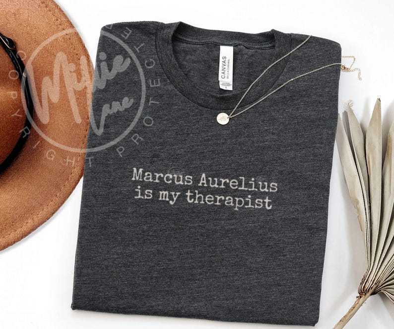 以下が含まれることがあります： 「Marcus Aurelius is my therapist」の白文字がプリントされた濃いグレーのTシャツ。丸いペンダントが付いたゴールドカラーのネックレスがシャツの上にあります。茶色の帽子が一部見えています。