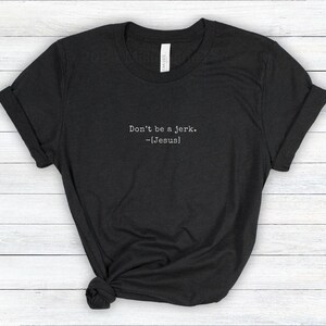 Peut inclure: Un t-shirt gris fonc&eacute; avec le texte "Don't be a jerk. -[Jesus]" imprim&eacute; en blanc. Le t-shirt est pli&eacute; et pos&eacute; sur une surface claire &agrave; grain de bois.