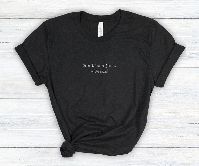 Peut inclure: Un t-shirt gris fonc&eacute; avec le texte "Don't be a jerk. -[Jesus]" imprim&eacute; en blanc. Le t-shirt est pli&eacute; et pos&eacute; sur une surface claire &agrave; grain de bois.