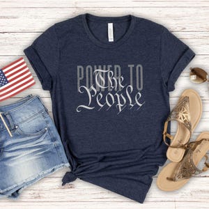 May include: A navy blue t-shirt with the text "Power to the People" printed in white. The text is stylized to look like the preamble to the US Constitution. The shirt is laid out on a white wooden surface with a pair of denim shorts, a small American flag, and two pairs of sandals.