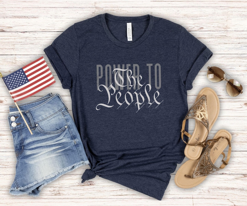 May include: A navy blue t-shirt with the text "Power to the People" printed in white. The text is stylized to look like the preamble to the US Constitution. The shirt is laid out on a white wooden surface with a pair of denim shorts, a small American flag, and two pairs of sandals.