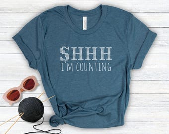 May include: A heathered blue short-sleeve t-shirt featuring the white text SHHH I'M COUNTING, with the word SHHH rendered in a knit-texture font. Beside the shirt are pink-framed sunglasses and a ball of black yarn with knitting needles.