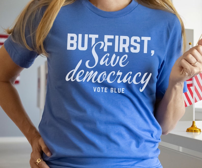 May include: A blue t-shirt with the text "BUT FIRST, Save democracy VOTE BLUE" in white. The shirt is a solid color and has short sleeves. The person is holding a small American flag.