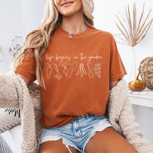 May include: A woman wearing a rust-coloured t-shirt with the text "life begins in the garden" and a white outline of gardening tools and vegetables. She is wearing denim shorts and a white fluffy cardigan.