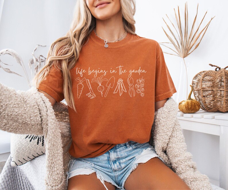 May include: A woman wearing a rust-coloured t-shirt with the text "life begins in the garden" and a white outline of gardening tools and vegetables. She is wearing denim shorts and a white fluffy cardigan.