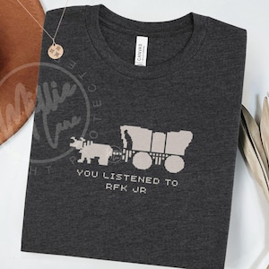 Puede incluir: Camiseta gris oscuro con un oso pixelado tirando de una carreta cubierta. El texto "YOU LISTENED TO RFK JR" est&aacute; debajo del gr&aacute;fico. La camiseta est&aacute; hecha de un material suave.