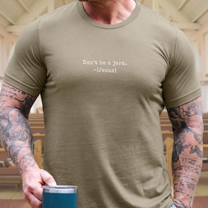 Peut inclure: T-shirt vert olive avec le texte "Don't be a jerk. -[Jesus]" en blanc. Le t-shirt est &agrave; manches courtes et a un col rond. La personne portant le t-shirt a des tatouages sur les bras.