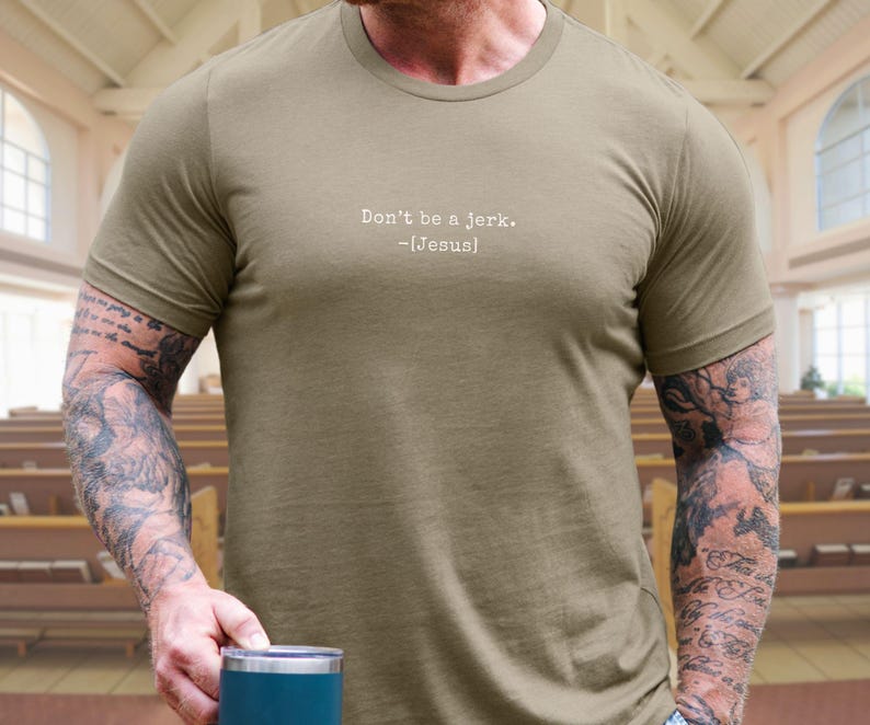 Peut inclure: T-shirt vert olive avec le texte "Don't be a jerk. -[Jesus]" en blanc. Le t-shirt est &agrave; manches courtes et a un col rond. La personne portant le t-shirt a des tatouages sur les bras.