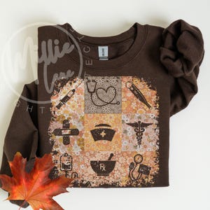 Peut inclure: Un sweat-shirt marron avec un motif floral et médical. Le motif présente un collage de symboles médicaux, dont un stéthoscope, une croix et un mortier et un pilon. Une feuille d'automne est en bas à gauche.