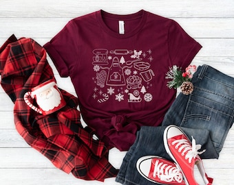 Könnte beinhalten: Ein maroonfarbenes T-Shirt mit einem weißen Design aus verschiedenen Weihnachtsback- und Feiertags-Icons. Das Design beinhaltet einen Nudelholz, eine Schüssel, ein Lebkuchenhaus, eine Schneeflocke, einen Zuckerstock, einen Weihnachtsbaum, einen Schlitten des Weihnachtsmanns und eine Schürze.