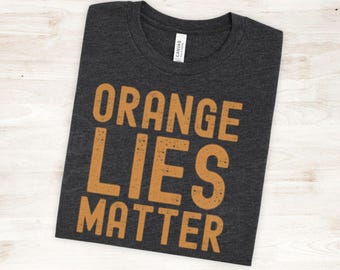 May include: Dark gray t-shirt with the text "ORANGE LIES MATTER" in orange block letters. The shirt is folded neatly, showcasing the bold statement. The shirt is made of a soft material.