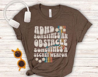May include: Brown t-shirt with a retro-style graphic that reads "ADHD Sometimes an Obstacle Sometimes a Secret Weapon". The graphic features colorful stripes and flowers.