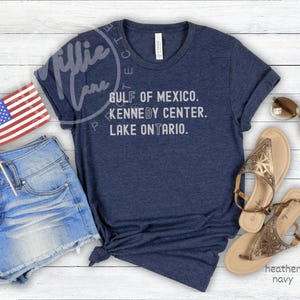 May include: A heather navy blue t-shirt featuring the white text GULF OF MEXICO. KENNEDY CENTER. LAKE ONTARIO. accompanied by blue denim shorts, a small American flag on a stick, a pair of gold metallic sandals, and brown-tinted sunglasses.