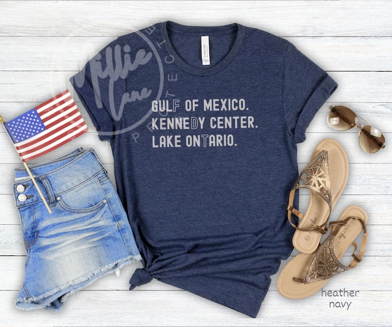 May include: A heather navy blue t-shirt featuring the white text GULF OF MEXICO. KENNEDY CENTER. LAKE ONTARIO. accompanied by blue denim shorts, a small American flag on a stick, a pair of gold metallic sandals, and brown-tinted sunglasses.
