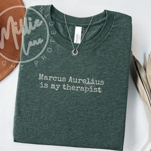 以下が含まれることがあります： 「Marcus Aurelius is my therapist」の白文字が入った濃い緑色のTシャツ。太陽のペンダントが付いたシルバーのネックレスがシャツの上にあります。シャツは白い表面に折りたたまれています。