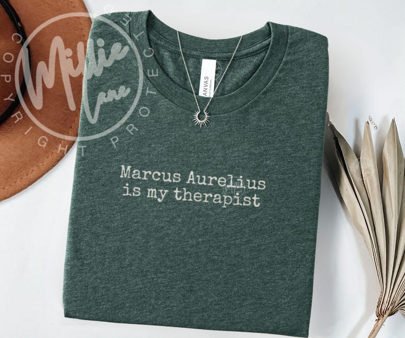 以下が含まれることがあります： 「Marcus Aurelius is my therapist」の白文字が入った濃い緑色のTシャツ。太陽のペンダントが付いたシルバーのネックレスがシャツの上にあります。シャツは白い表面に折りたたまれています。