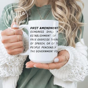 May include: A white ceramic mug featuring black printed text that reads: FIRST AMENDMENT CONGRESS SHALL MAKE NO LAW RESPECTING AN ESTABLISHMENT OF RELIGION, OR PROHIBITING THE FREE EXERCISE THEREOF; OR ABRIDGING THE FREEDOM OF SPEECH, OR OF THE PRESS; OR THE RIGHT OF THE PEOPLE PEACEABLY TO ASSEMBLE, AND TO PETITION THE GOVERNMENT FOR A REDRESS OF GRIEVANCES.
