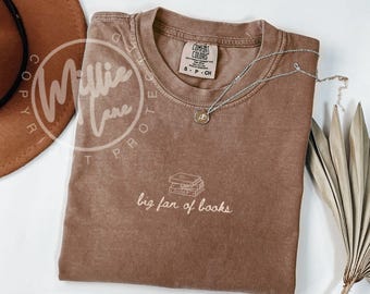 Peut inclure: Un t-shirt marron avec le texte "big fan of books" et un dessin d'une pile de livres. Un collier argenté avec un pendentif est sur le t-shirt. Un chapeau marron et une plante séchée sont en arrière-plan.