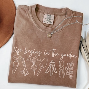 May include: A brown t-shirt with the text "life begins in the garden" printed in white. The shirt features a line drawing of gardening tools and vegetables, including a carrot, a rake, a beet, a trowel, a pair of pruning shears, and a tomato plant.
