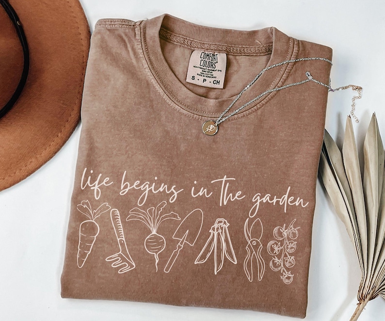 May include: A brown t-shirt with the text "life begins in the garden" printed in white. The shirt features a line drawing of gardening tools and vegetables, including a carrot, a rake, a beet, a trowel, a pair of pruning shears, and a tomato plant.