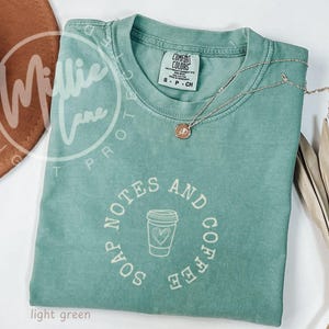 May include: A light green short-sleeve t-shirt featuring a circular graphic with the text "SOAP NOTES AND COFFEE" surrounding a line-art illustration of a coffee cup with a heart. The shirt has a crew neckline and a Comfort Colors brand label.