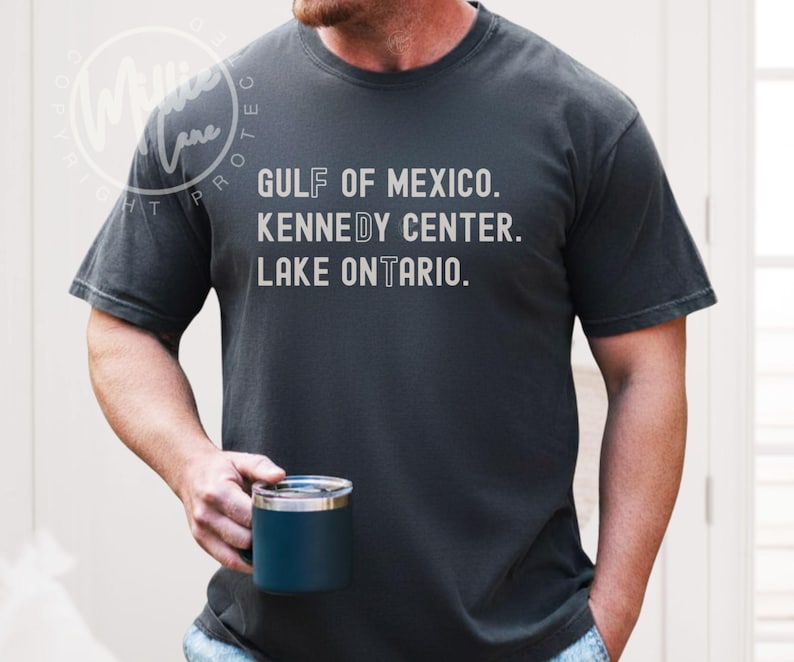 May include: A charcoal gray short-sleeve t-shirt featuring white text that reads GULF OF MEXICO, KENNEDY CENTER, and LAKE ONTARIO. The letters F, D, and T are highlighted in a different font style within the words on the shirt.