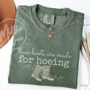Puede incluir: Camiseta verde musgo con el texto "These boots are made for hoeing" y un gráfico de dos botas. Un collar dorado con un colgante está en la camisa. La camiseta es de la marca Comfort Colors.