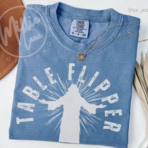 May include: A blue jean colored short-sleeve t-shirt featuring a white graphic print of a figure with arms outstretched and the text "TABLE FLIPPER" arched above. The shirt has a crew neckline and a Comfort Colors brand label at the inner collar.