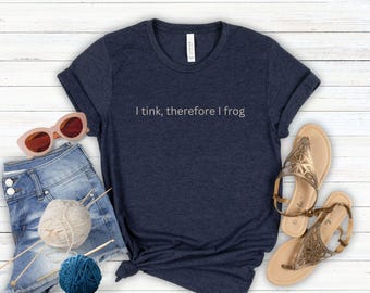 May include: A dark navy blue short-sleeved t-shirt with the text I tink, therefore I frog printed in white on the chest. The shirt is displayed alongside light blue denim shorts, a ball of beige yarn, a ball of blue yarn, knitting needles, sunglasses, and sandals.