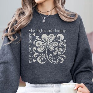 May include: A dark gray sweatshirt with a white graphic of a stylized four-leaf clover with the text "may your heart be light and happy".