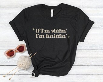 May include: A black short-sleeved t-shirt featuring the beige text "if I'm sittin' I'm knittin'" with small star accents. The shirt is displayed alongside a ball of light-colored yarn with two knitting needles and a pair of pink-framed sunglasses.