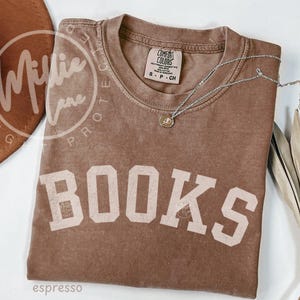 May include: A brown short-sleeve t-shirt with the word BOOKS printed in large, off-white block letters across the chest. The shirt features a Comfort Colors label at the inner neckline, indicating size S, and is paired with a thin silver necklace.
