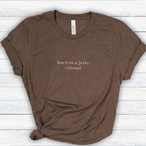 Peut inclure: T-shirt marron avec des manches roul&eacute;es. Le t-shirt porte l'inscription "Don't be a jerk. -[Jesus]" dans une police simple sans empattement. Le t-shirt est fait d'une mati&egrave;re douce et est pr&eacute;sent&eacute; sur une surface claire en grain de bois.
