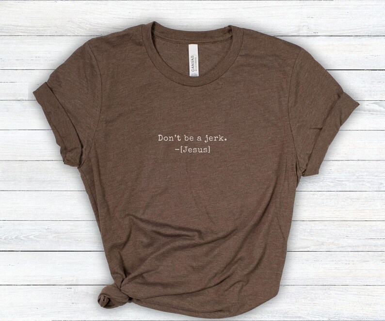 Peut inclure: T-shirt marron avec des manches roul&eacute;es. Le t-shirt porte l'inscription "Don't be a jerk. -[Jesus]" dans une police simple sans empattement. Le t-shirt est fait d'une mati&egrave;re douce et est pr&eacute;sent&eacute; sur une surface claire en grain de bois.