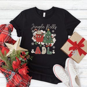 May include: A black short-sleeve t-shirt featuring a graphic of a red mug and a green Christmas tree with the text Jingle Bells & ADLs. The design includes white snowflakes and cartoon-style faces on the mug and tree, with red and green lettering.
