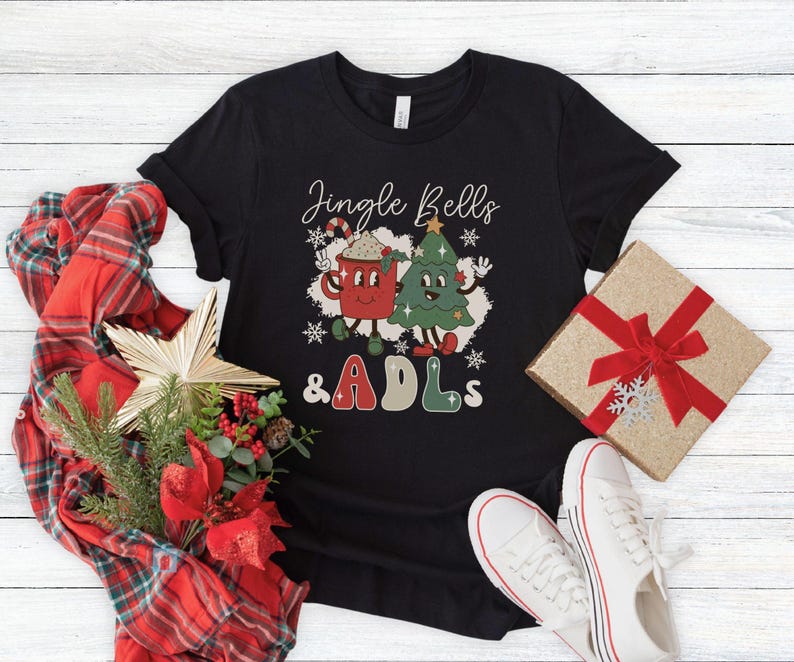 May include: A black short-sleeve t-shirt featuring a graphic of a red mug and a green Christmas tree with the text Jingle Bells & ADLs. The design includes white snowflakes and cartoon-style faces on the mug and tree, with red and green lettering.