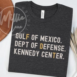 May include: Dark grey t-shirt with white text: "GULF OF MEXICO. DEPT OF DEFENSE. KENNEDY CENTER." The word "DEFENSE" has a yellow "D" and "T" in "CENTER" is also yellow. A white tag reads "CANVAS".