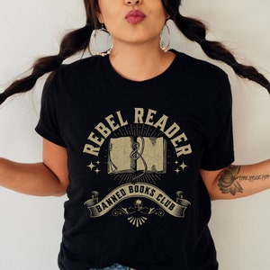 K&ouml;nnte beinhalten: Ein schwarzes T-Shirt mit einem Vintage-Grafikdesign, auf dem "Rebel Reader Banned Books Club" steht. Die Grafik zeigt ein Buch mit einer Schlange, die sich darum windet, Sterne und ein Banner.