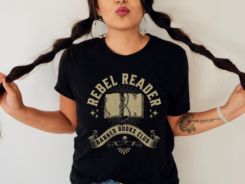 K&ouml;nnte beinhalten: Ein schwarzes T-Shirt mit einem Vintage-Grafikdesign, auf dem "Rebel Reader Banned Books Club" steht. Die Grafik zeigt ein Buch mit einer Schlange, die sich darum windet, Sterne und ein Banner.