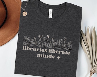 May include: A dark gray t-shirt with a graphic of books, flowers, and stars. The text "libraries liberate minds" is printed in a light beige color on the shirt.