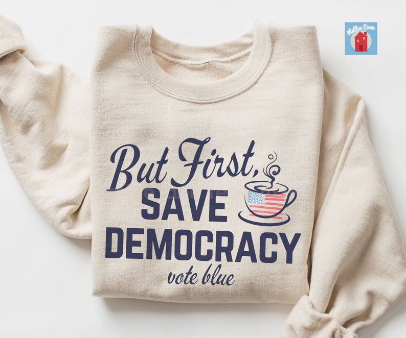 May include: A beige sweatshirt with the text "But First, Save Democracy vote blue" in navy blue lettering. A coffee cup with an American flag design is above the text.
