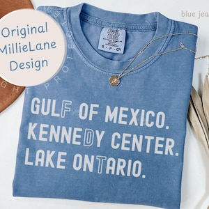May include: A blue jean colored t-shirt featuring white text that reads GULF OF MEXICO. KENNEDY CENTER. LAKE ONTARIO. The shirt has a crew neckline and a Comfort Colors brand label on the inside collar. A thin gold chain necklace rests on the fabric.