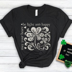May include: A black t-shirt with a white graphic of a four-leaf clover with a decorative swirl design. The text "be light and happy may your heart" is printed on the shirt.