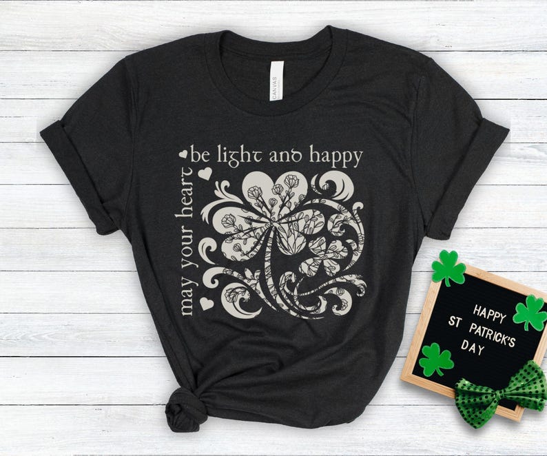 May include: A black t-shirt with a white graphic of a four-leaf clover with a decorative swirl design. The text "be light and happy may your heart" is printed on the shirt.